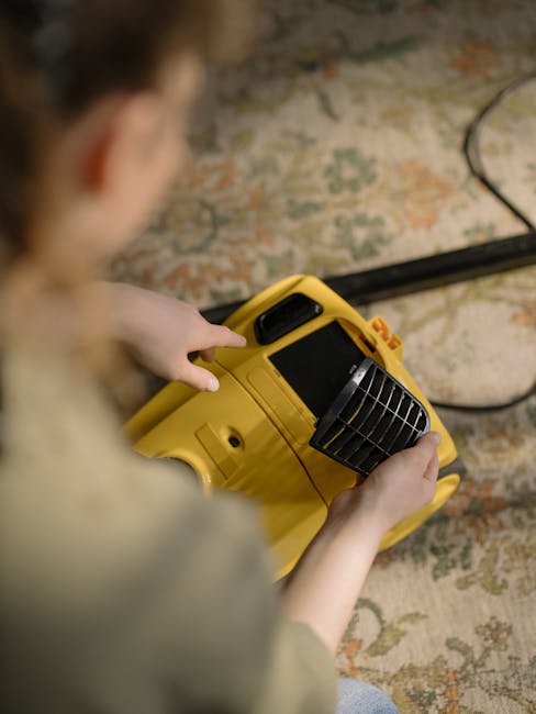 A person holding a yellow portable carpet cleaning machine with a black vent, preparing for surface cleaning or deep cleaning in an indoor setting. The background features a patterned carpet with mute