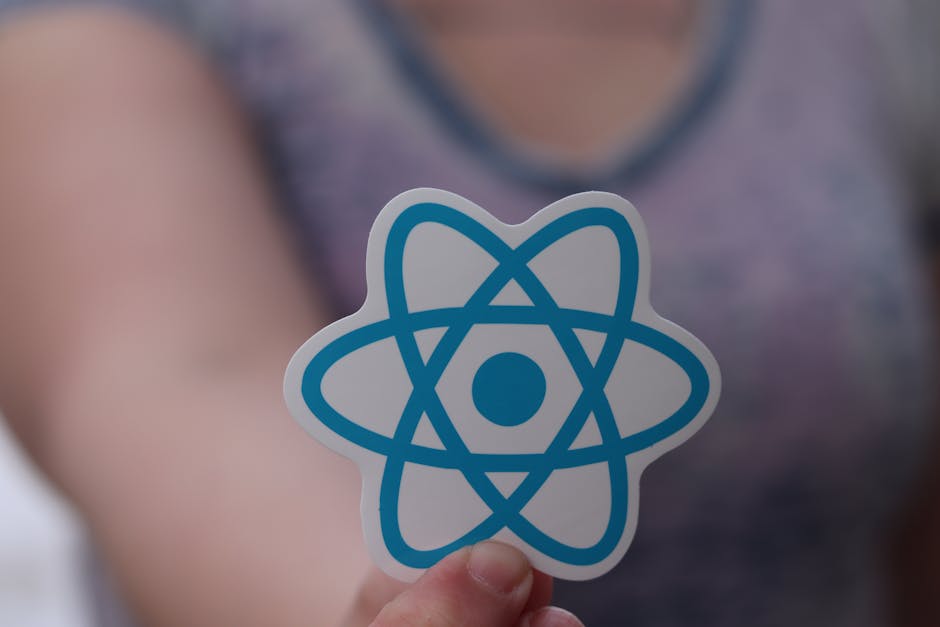 A person holding a paper cutout of a blue atomic model symbol in front of their torso, which is blurred and dressed in a purple top, inside a room with soft, diffused lighting; the focus is on the ato