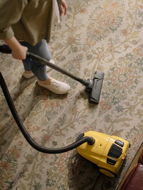 A person kneeling on a patterned carpeted floor, using a handheld cloth to clean the interior of a yellow and black vacuum cleaner with a flexible hose. The room appears well-lit, with the carpet's de